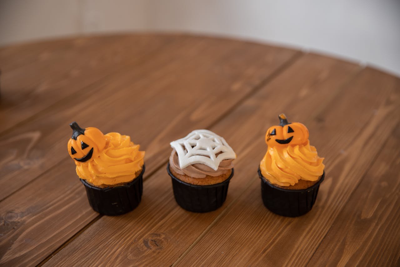 Three Halloween-themed cupcakes with pumpkin and spider web designs on a wooden table.